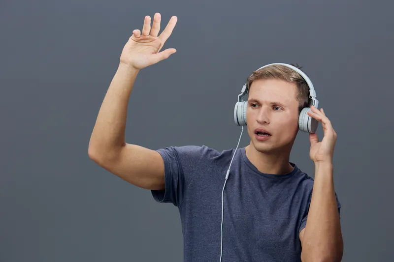 Young man with headphones and casual clothing gesturing with his hand while talking or listening, isolated on plain gray background. Modern lifestyle, technology, communication concept.