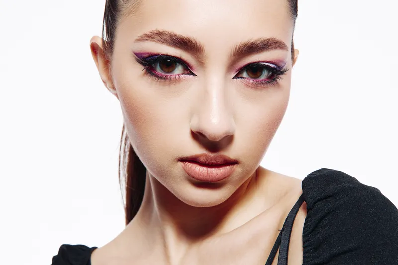 Closeup portrait of a stylish young woman with creative pink eye makeup and natural matte lips, wearing a black top on white background. Concept of fashion beauty makeup artistry.