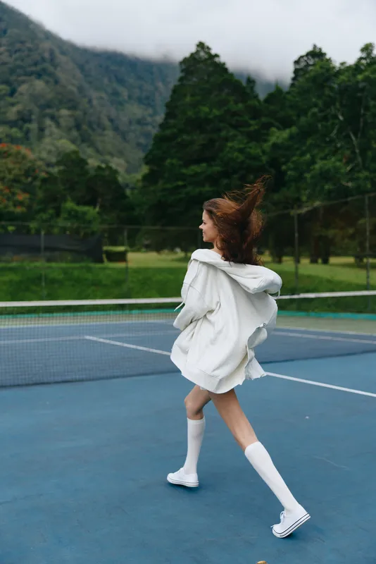Young woman in sporty attire joyfully running on a tennis court, surrounded by lush greenery and mountains, conveying a sense of energy and vitality in a refreshing setting