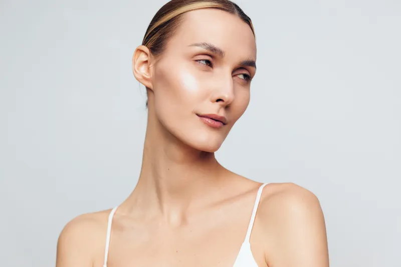 Portrait of a woman with clear skin looking sideways, soft natural light, minimal makeup, neutral background, elegant and calm expression, simple white top visible