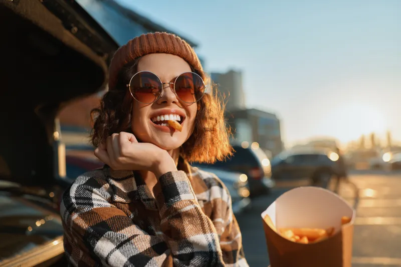 woman smile fries sunglasses beanie golden hour candid urban portrait of a joyful young woman eating street fries at sunset, authenticity and candid lifestyle with golden hour glow, mindful living