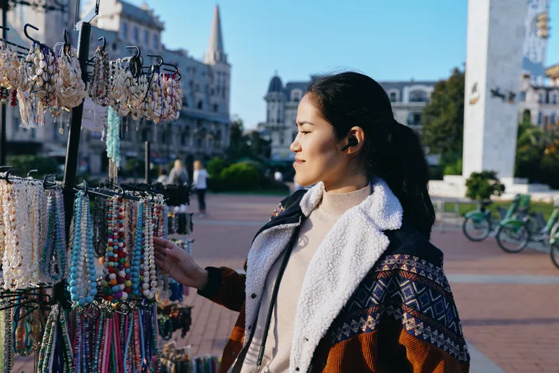 Woman admires colorful bead jewelry at an outdoor market stall, browsing necklaces and bracelets as sunlight lights the plaza, showcasing craft and fashion in a lively street scene
