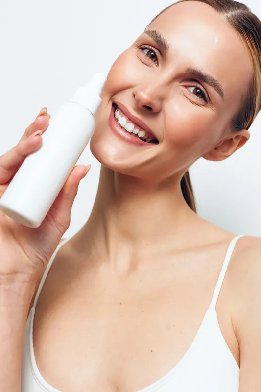 Smiling young woman with clear skin holds white cosmetic bottle close to face, shows natural beauty and skincare routine, isolated on white background with positive expression.