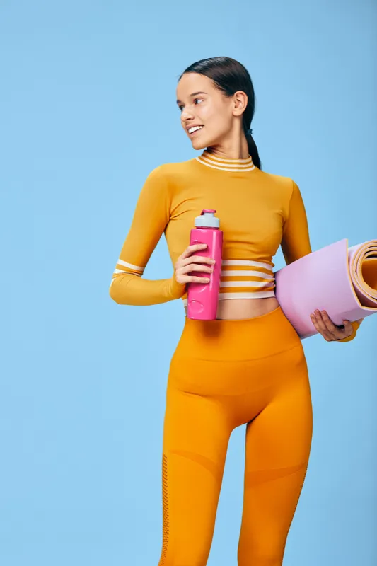 Woman fitness sport portrait: fit woman holding pink water bottle and rolled purple yoga mat, wearing bright orange athletic leggings and crop top, smiling with confident posture, ready for workout