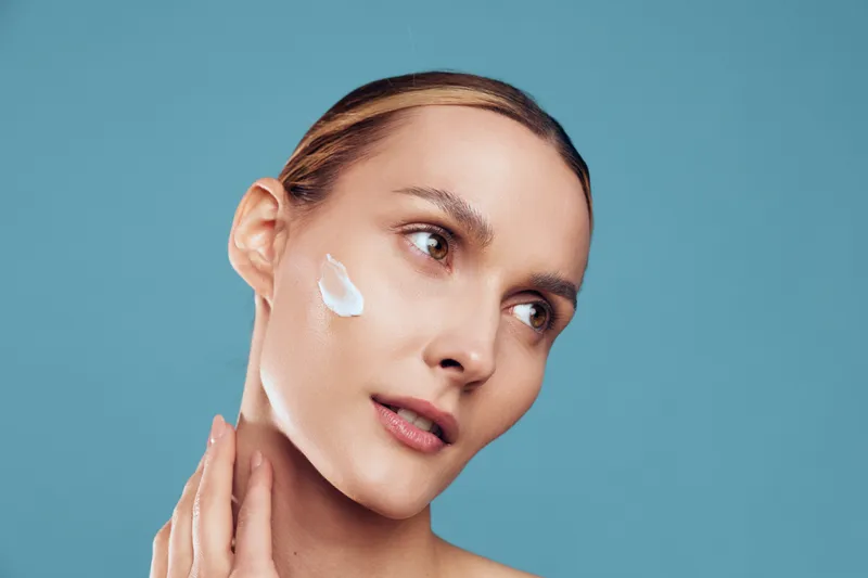 Woman skincare face close up with cream and serum on cheek, beauty model touching jaw, calm look, smooth hydrated skin, gentle wellness mood, isolated on solid blue background.