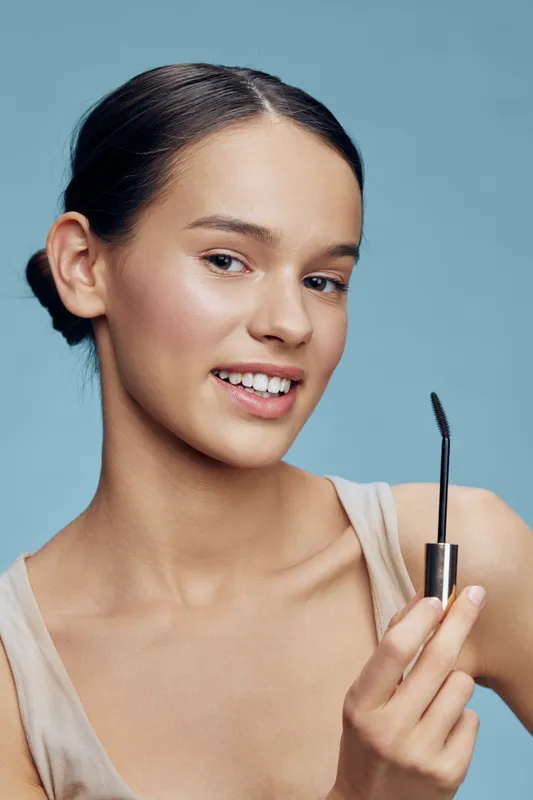 Young woman applying mascara with a brush on a blue background, highlighting beauty and makeup trends in a modern and fresh style