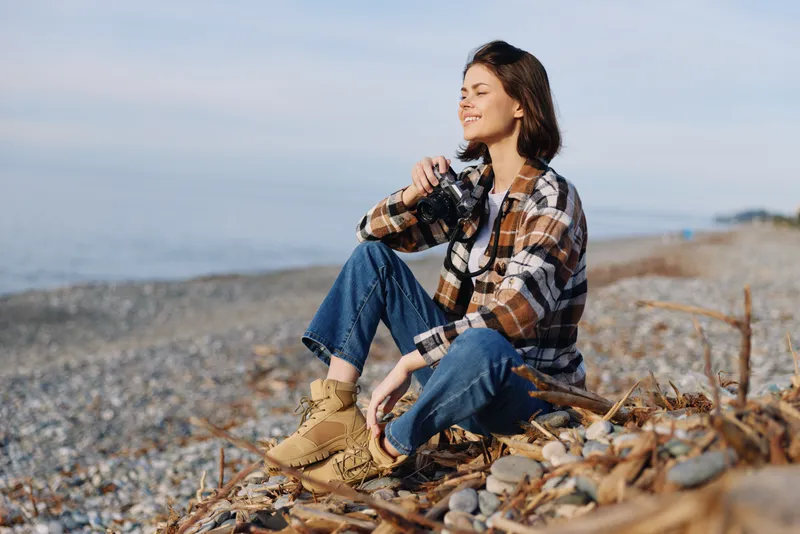 Woman photographer sits on a rocky beach with a camera hanging from her neck, wearing a plaid shirt and denim, enjoying outdoor scenery and sea breeze in a peaceful landscape.