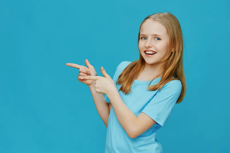 Happy girl in a light blue shirt pointing with smiles on a bright blue background, emphasizing joy and playful energy Perfect for children s clothing and lifestyle concepts