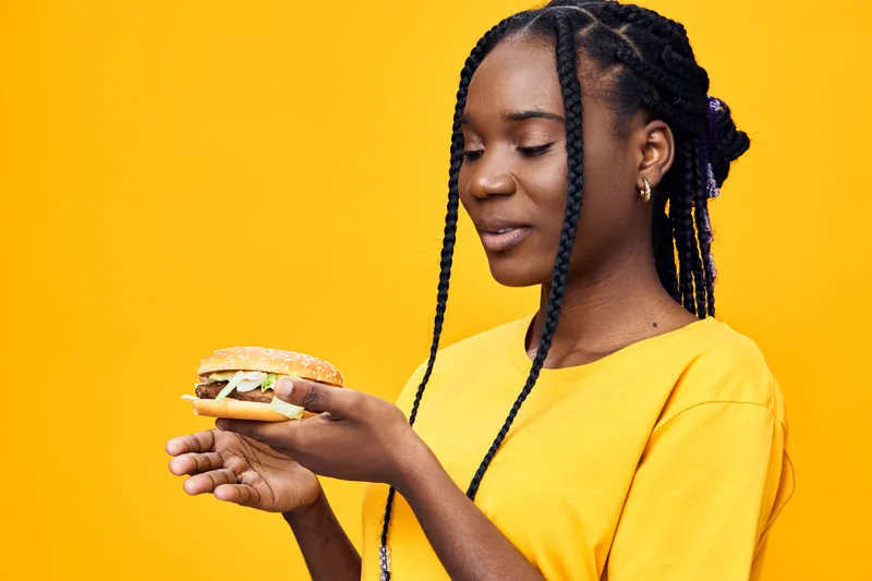 Young Black woman holding a delicious burger, showcasing vibrant yellow background and casual style, conveying happiness and enjoyment of healthy eating