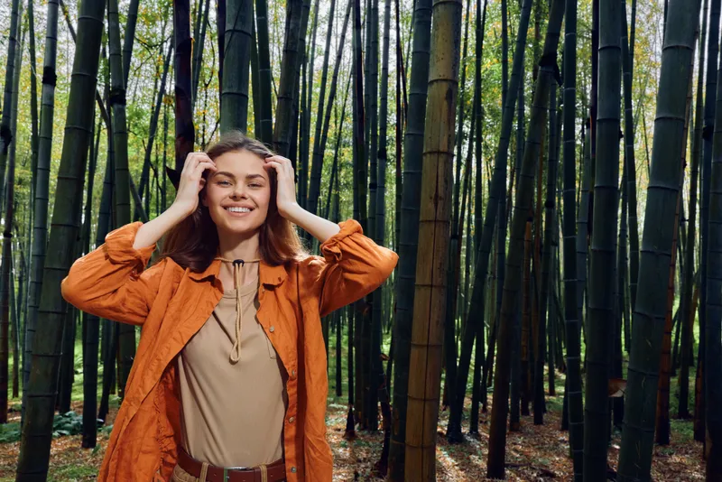 Woman in orange jacket stands smiling in bamboo forest, smile and relaxed portrait in nature. Outdoor scene with tall trees, green stalks, soft sunlight and serene atmosphere.