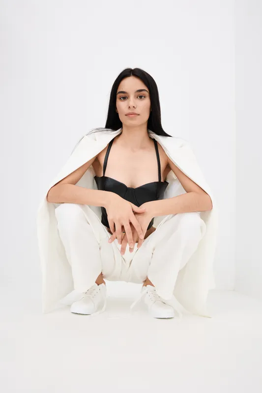 Young woman squatting in minimalist white studio wearing a white suit jacket, white pants, black top, and white sneakers with a confident, calm expression.
