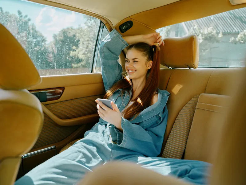 Young woman in a denim outfit smiles while using her phone in a luxury car interior, showcasing a casual and joyful mood in a warm, sunny atmosphere.