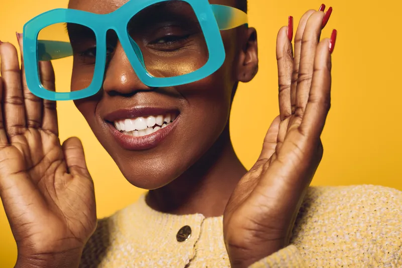 Happy African woman with dark skin wearing oversized blue glasses and a yellow sweater smiles brightly against yellow background. Concept of joy, fashion, and vibrant lifestyle.