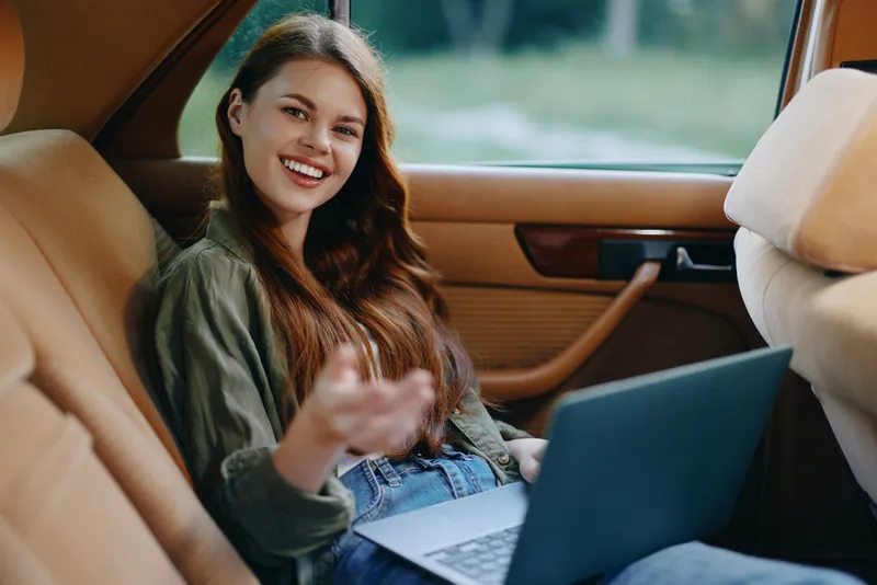 Young woman with long hair smiling and using a laptop in the backseat of a car on a sunny day, showcasing comfort and modern lifestyle.