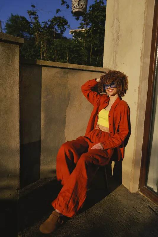 Young woman wearing a bright orange suit sits on a sunlit balcony, striking a relaxed pose with a casual smile and confident aura