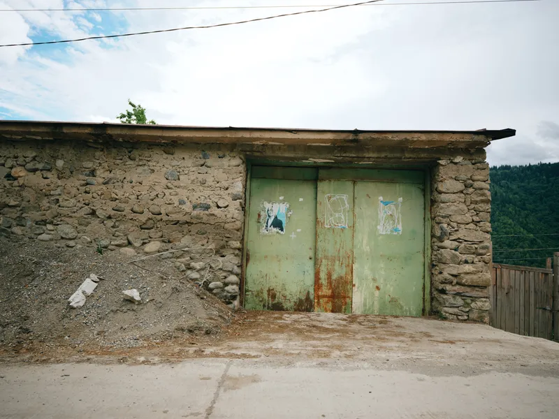 Rustic stone building with weathered green metal doors, outdoor scene with peeling posters, rough wall, rural industrial vibe, crumbling masonry and worn textures. With mellow light. Soft.