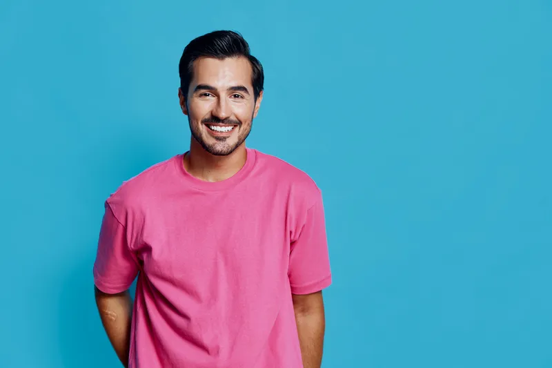 Smiling young man wearing pink t-shirt stands against a vibrant blue background, radiating positivity and friendliness in a studio portrait setting.