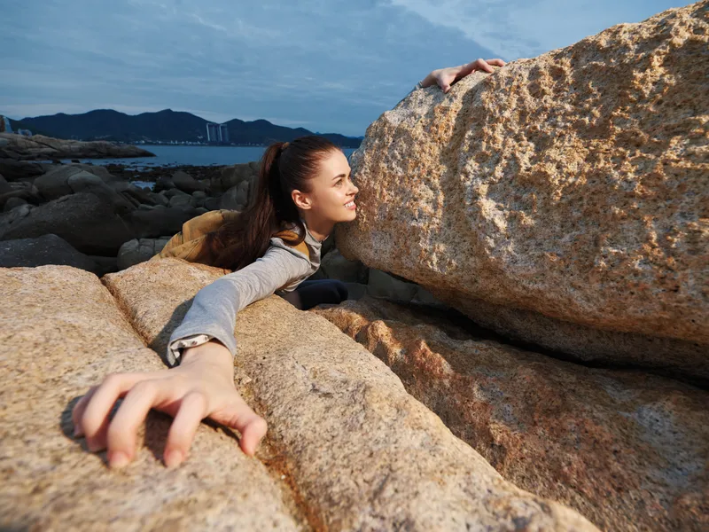 Adventurous young woman climbing rocky terrain, showcasing determination and strength against a coastal backdrop with beautiful natural lighting