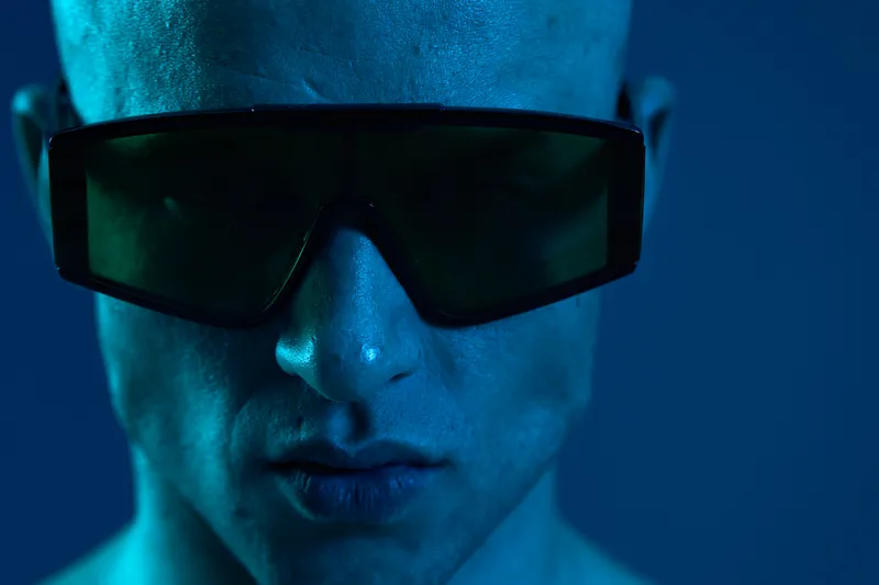 Close-up of a man wearing sunglasses, showcasing striking expression in a cool blue tone, emphasizing strong features and contemporary style.