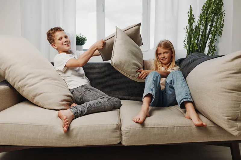 Two children having fun on beige sofa in bright living room with pillows in hands. Concept of playful siblings, childhood joy, and family bonding at home.