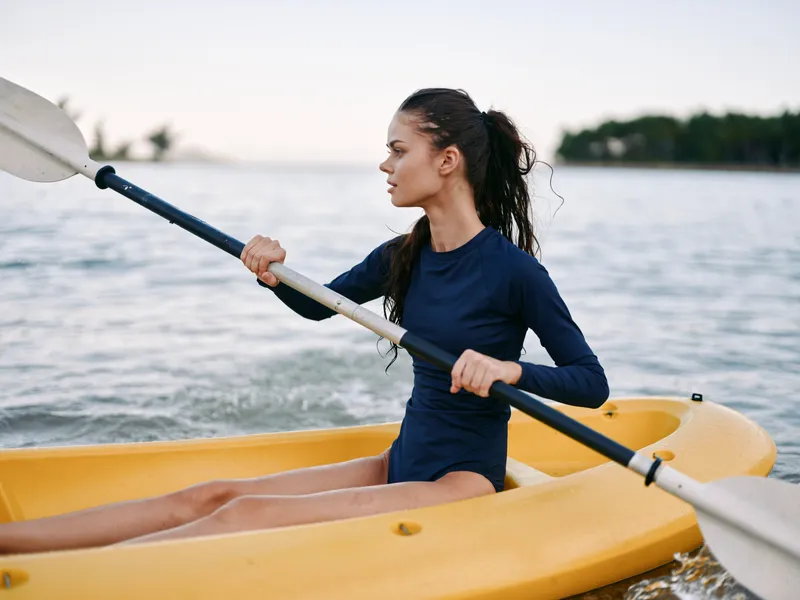 Female kayaker paddling a yellow kayak on calm water, wearing a dark blue wetsuit, focused expression as oar blades slice through gentle ripples and wind.