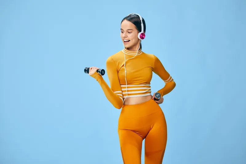 Energetic woman fitness model in orange athletic wear lifting dumbbells with headphones against a bright blue studio backdrop, capturing movement, balance and motivation during a dynamic workout.