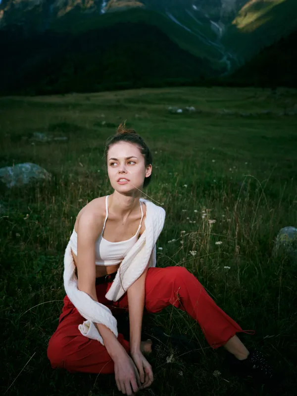 Woman sits in a grassy field with distant mountains, wearing a white top, red pants and a cardigan, capturing relaxed outdoor style and natural scenery