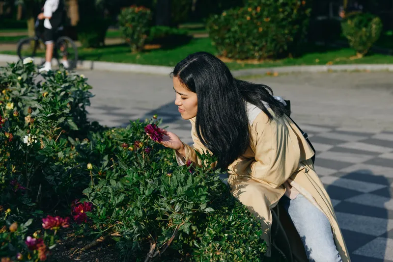 Woman leaning close to flowering bushes in a park, smelling blooms, wearing a beige coat and dark hair, sunlight and greenery framing a peaceful outdoor moment of natural beauty and floral exploration