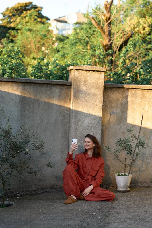 A woman wearing a red jumpsuit sits on the ground outdoors, taking a selfie with a phone against a weathered wall, showcasing casual fashion, daily lifestyle, and a warm relaxed mood