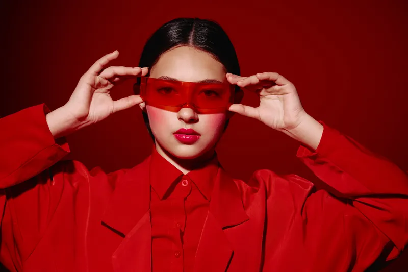 Young woman in vibrant red fashion holds transparent red sunglasses with both hands, wearing matching red outfit against radial red background. Concept of beauty, creative styling.