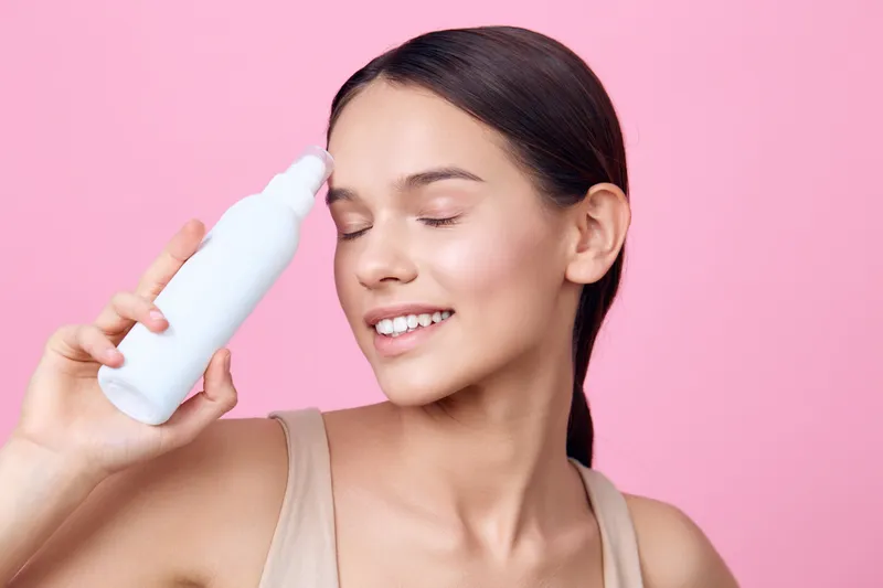 Young woman applying skincare product on her face with a joyful expression in a soft pink background, emphasizing beauty and self care routines