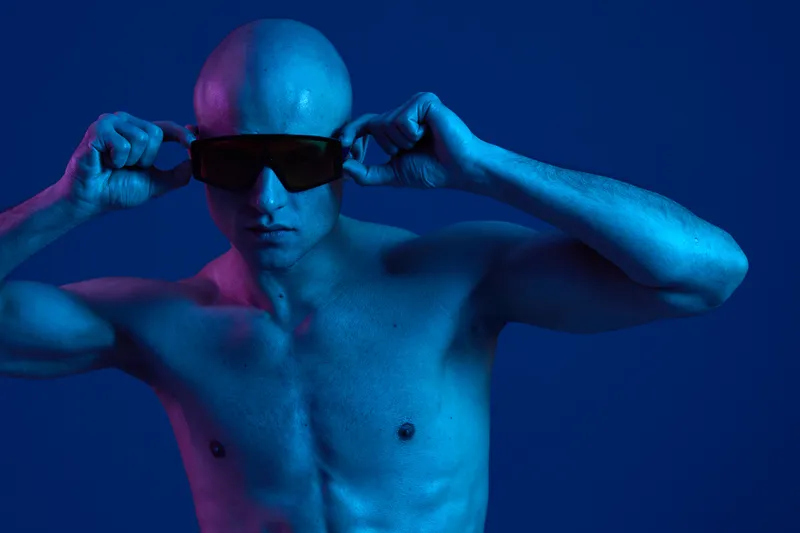 Athletic man in sunglasses poses confidently against a vibrant blue backdrop, showcasing a striking contrast with the lighting, enhancing the visual impact.