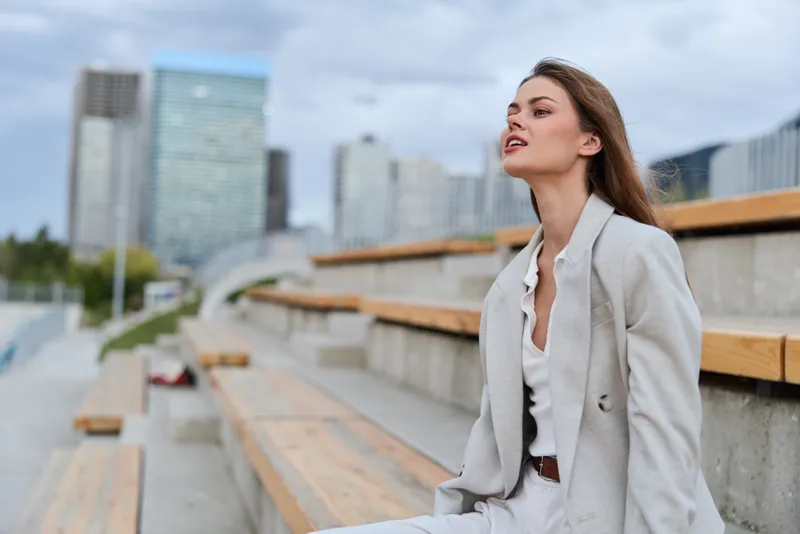 Woman in light gray suit feeling thoughtful in urban outdoor seating with modern city skyline background, business style and autumn mood