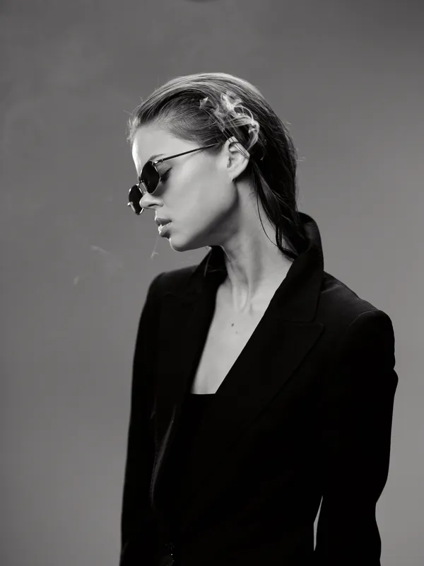 Black and white portrait of a stylish woman in sunglasses wearing a black blazer with a serious expression.