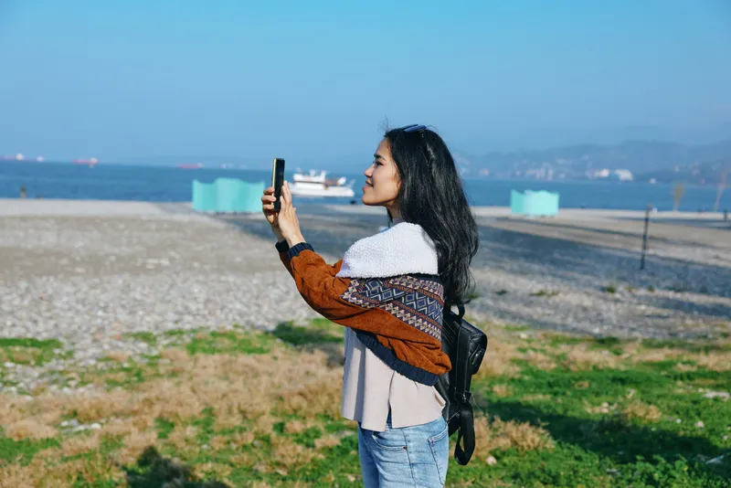 Woman capturing seaside scenery with smartphone on a sunny beach, exploring travel moments, casual style, knit shawl and backpack, blue water and expansive horizon