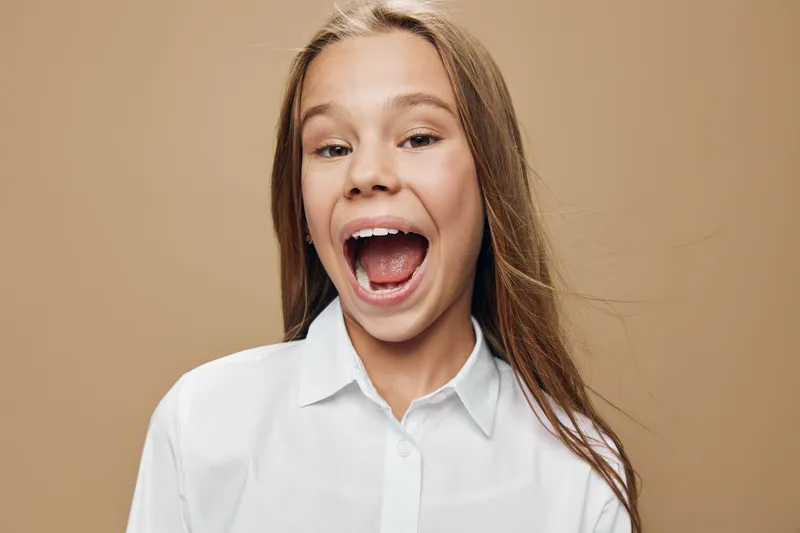 Happy teenage girl with long hair wearing white shirt smiling and laughing on beige background. Youthful female portrait with open mouth expressing joy and excitement in studio.