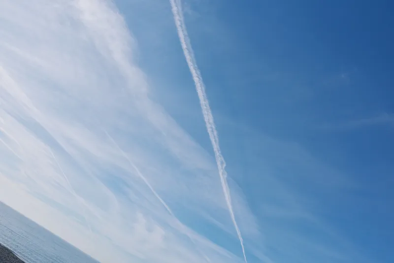 Blue sky above calm water with a distant horizon, a single contrail diagonally crosses the scene, evoking travel, weather and expansive outdoor serenity.