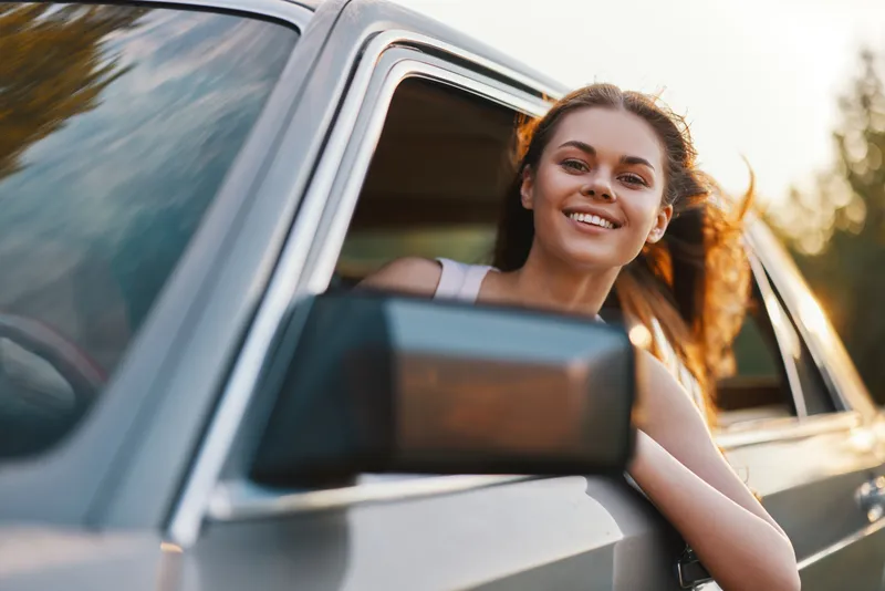 Happy young woman enjoying a sunny day driving in a classic car, with flowing hair and vibrant smile, surrounded by a lush green background.