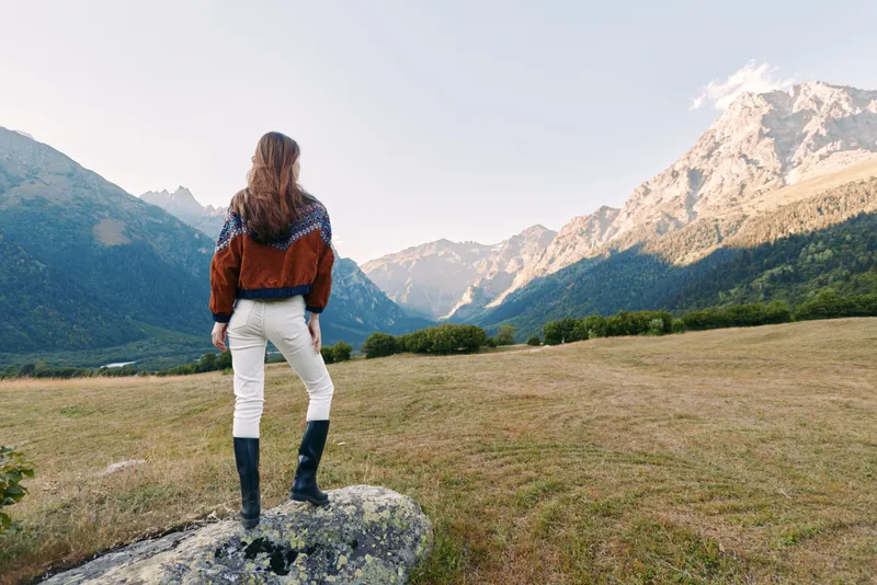 Woman standing on a rock overlooking mountains and a wide valley, nature landscape for hiking outdoors adventure, panoramic viewpoint with meadow, grass, alpine peak and sunlight.