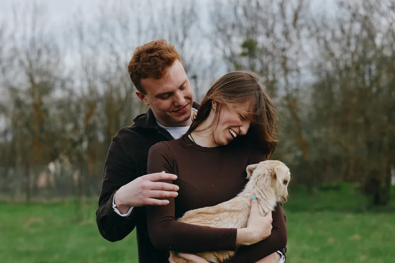 Couple outdoor goat kid laughter affection countryside nature breeze warmth as they smile and embrace in a sunlit moment, rustic setting, peaceful scene, natural colors, candid lifestyle