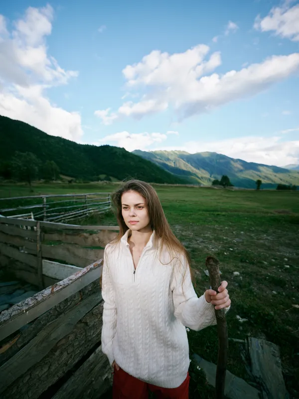 Rural countryside scene with a woman by a wooden fence holding a tool, open field and hills under a blue sky, capturing outdoor farming and nature in a calm landscape, light breeze