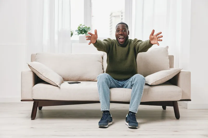 Happy African American man celebrating at home on a cozy sofa, wearing a casual sweater and jeans, expressing joy in a bright and inviting living room