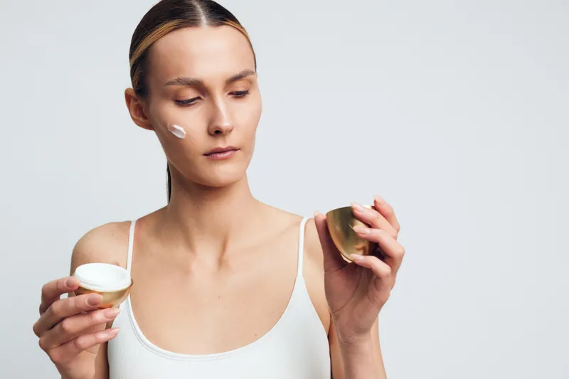 Woman applying face cream with cotton pad during skincare routine in a studio close up portrait soft light on neutral background shows self care wellness and beauty