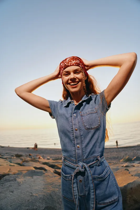 smiling woman in denim jumpsuit with boho-western red bandana enjoying lifestyle on beach at sunset with film color effect