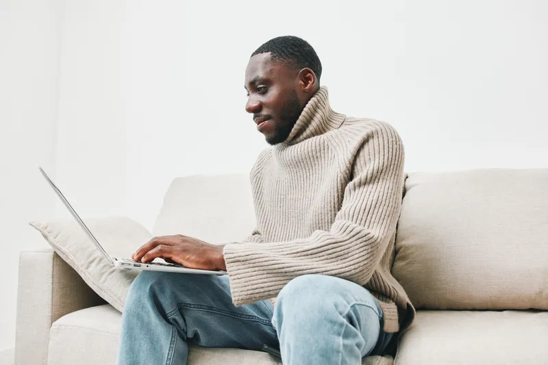 Young African man in cozy beige sweater working on laptop in bright living room, conveying focus and productivity, ideal for home office themes