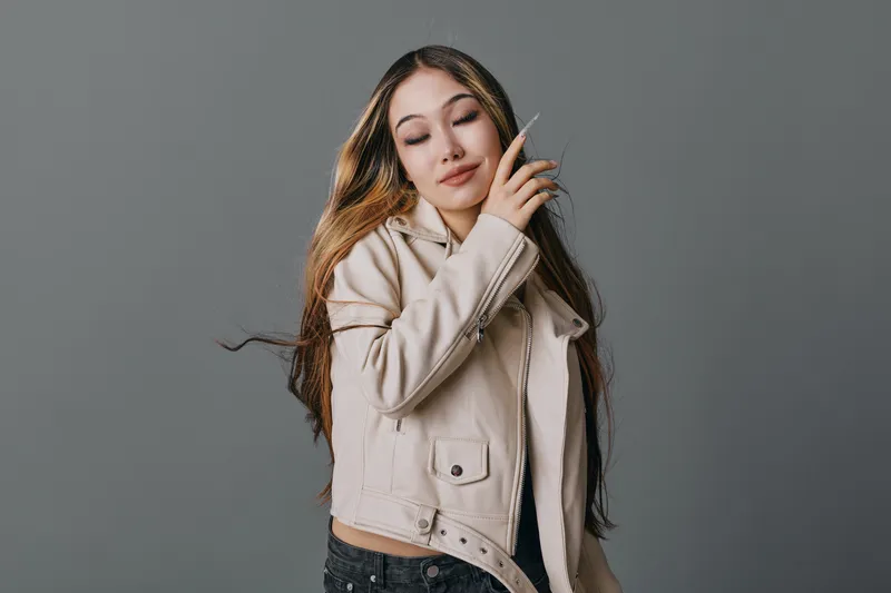 Young woman with long hair and beige jacket posing with eyes closed and hand near face on gray background. Fashion model showing relaxed expression and casual style indoor.
