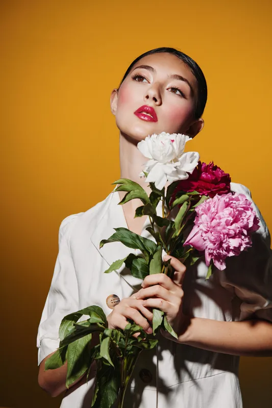 Creative young woman with glossy red lips and flawless makeup holds a bouquet of peonies against a warm yellow background. Concept of beauty, fashion, and elegance in modern portraits.