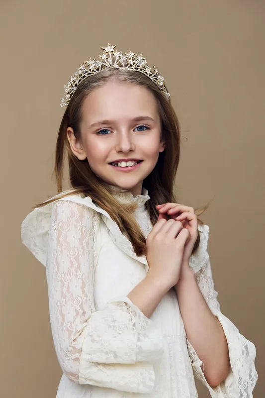Cheerful young girl in a elegant white dress with a tiara, smiling kindly against a soft beige background, showcasing innocence and joy