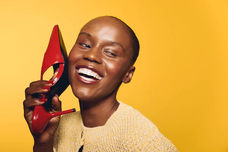 Happy African woman with dark skin holding red high heel shoe near her ear smiling on yellow background. Concept of joy, fashion, beauty, and positive emotions in studio lighting.