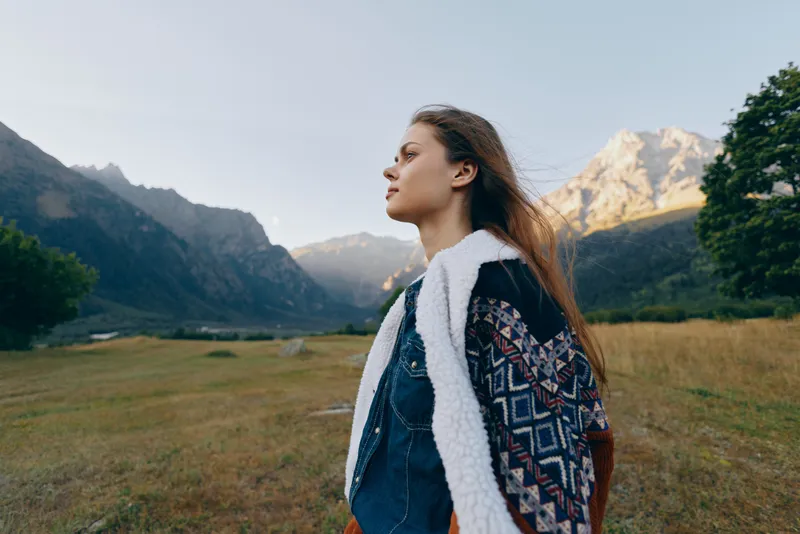 Woman profile in mountains valley nature portrait outdoors landscape wearing patterned jacket with shearling, contemplative young traveler gazing across alpine meadow in serene sunlight.